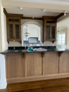 What Is the Average Cost of Custom Kitchen Cabinets? (2026 Guide) A kitchen with handcrafted solid wood cabinetry, a black countertop, and an open book and paint can on the counter. In the background, there is a white archway and a stove with a blue tile backsplash.