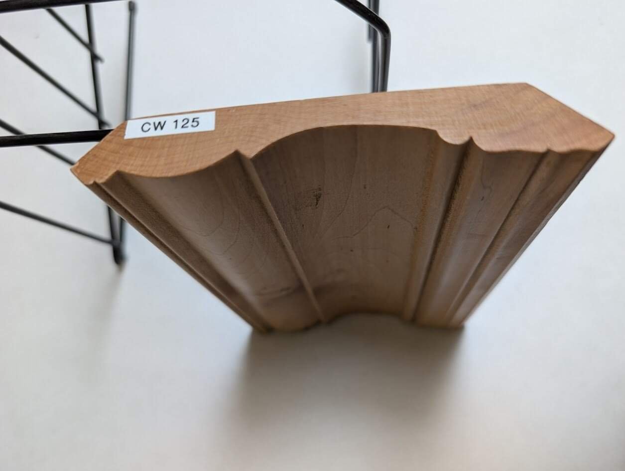 A piece of decorative wooden molding labeled CW 125 rests on a metal stand against a white background. Ideal for custom kitchen cabinets Pennsylvania, the molding features a smooth, curved profile with detailed edges.