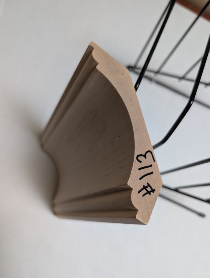 A piece of light wood molding with decorative edges, labeled #113 in black marker, stands upright on a white surface. Perfect for bespoke cabinetry manufacturers or cabinet makers in Honey Brook, PA; a wire rack is partially visible in the background.
