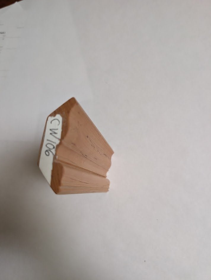 A small, triangular piece of wood trim with a white label marked CW06 sits on a white surface. The light brown wood, often found in handcrafted solid wood cabinetry, features angled edges and fine craftsmanship.