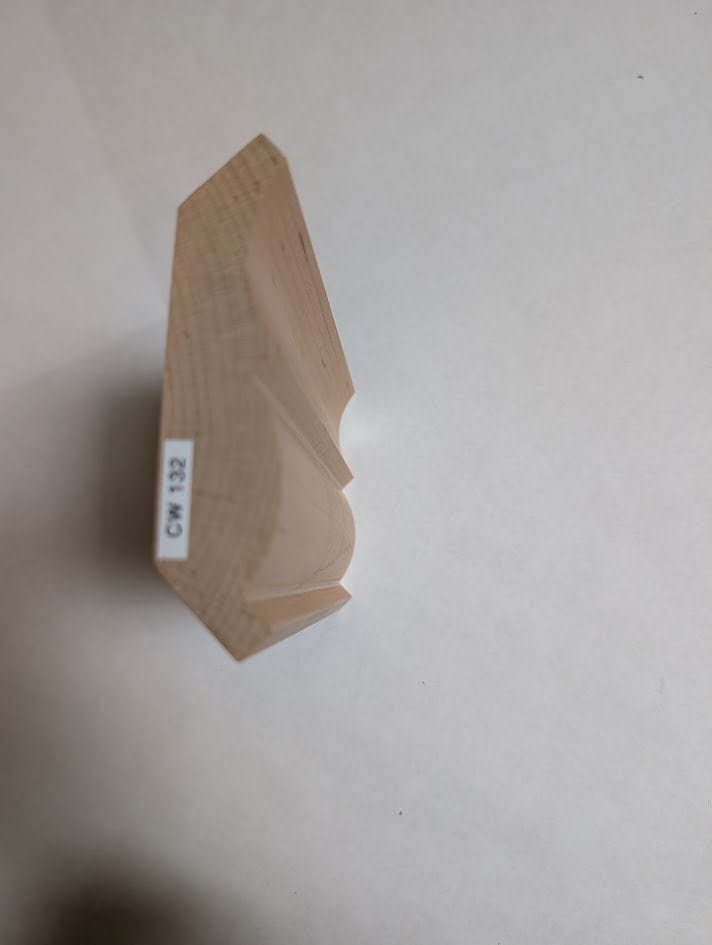 A piece of light-colored wood molding rests on a white surface, viewed from above at an angle. The molding has a decorative profile with grooves—ideal for custom kitchen cabinets Pennsylvania—and a sticker labeled CW-132 on one side.
