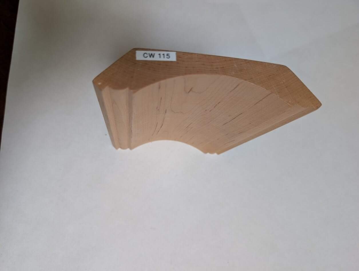 A wooden molding sample with a curved and angled profile, labeled CW 115, is placed on a white surface. The smooth finish and visible grain patterns highlight the quality found in handcrafted solid wood cabinetry.